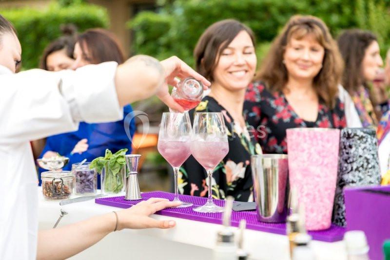 Un barman verse un liquide rose dans des verres à cocktail, devant un groupe de personnes souriantes en extérieur.