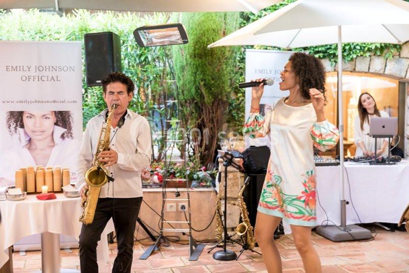 Un saxophoniste et une chanteuse se produisent en extérieur, avec une DJ en arrière-plan près d'une table.