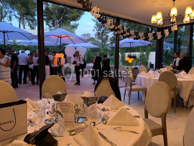 Salle de réception élégante avec tables dressées, vue sur une terrasse où des invités discutent sous des parasols.