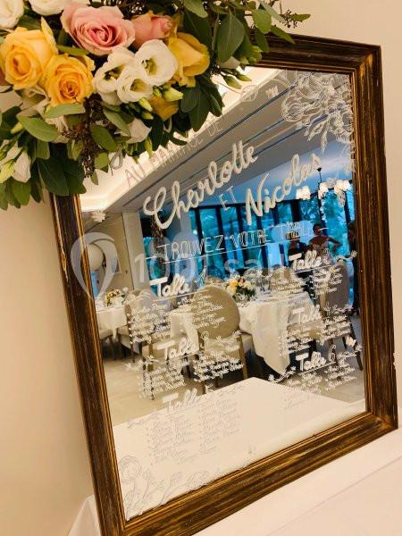 Plan de table écrit sur un miroir encadré, décoré de fleurs, avec une salle de réception visible en arrière-plan.