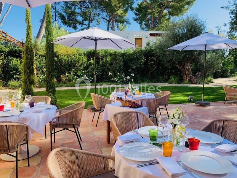 Terrasse ensoleillée avec tables dressées, chaises en rotin, parasols blancs et jardin verdoyant en arrière-plan.