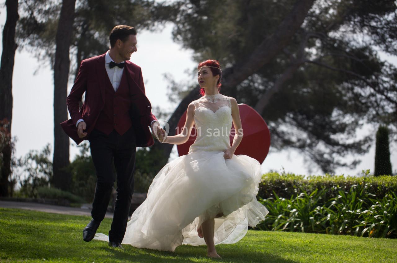 Un couple en tenue de mariage marche main dans la main sur une pelouse, entouré d'arbres et de verdure.