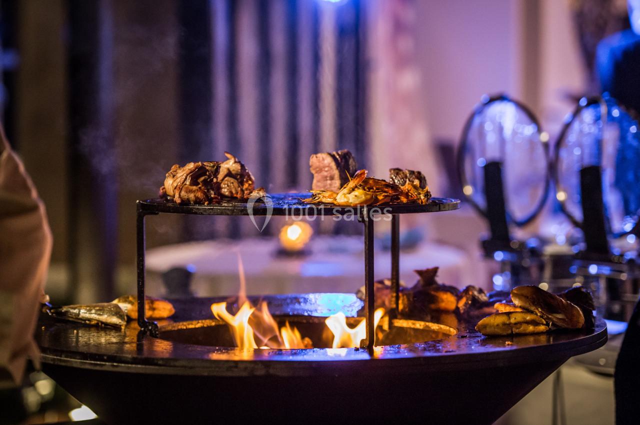 Viande et légumes grillant sur un brasero rond avec des flammes vives, dans un cadre chaleureux en soirée.