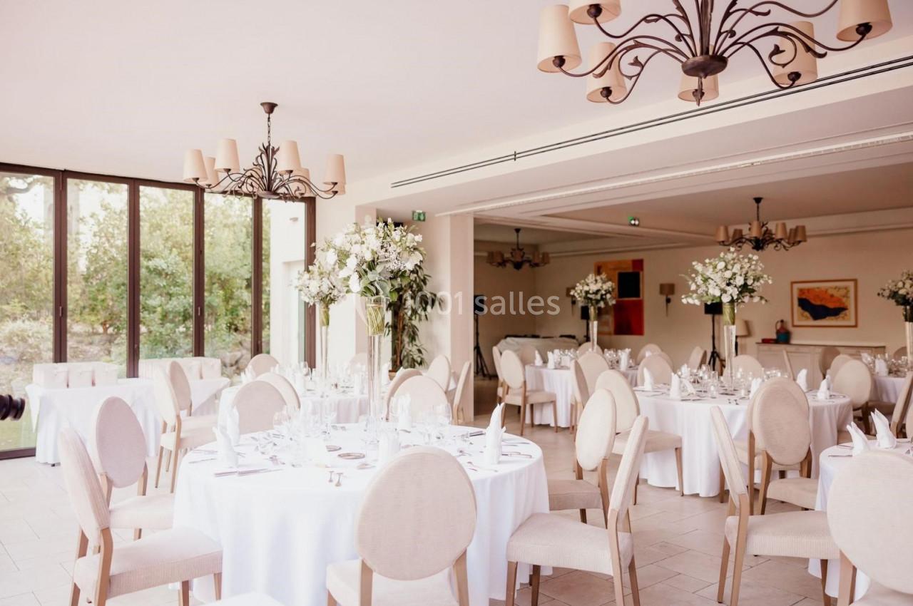 Salle de réception lumineuse avec tables rondes dressées, chaises beige clair et décorations florales blanches.