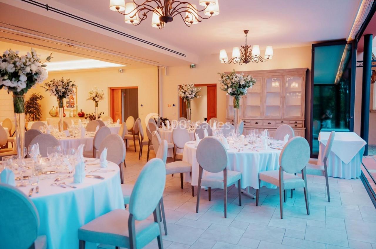 Salle de réception élégante avec tables rondes dressées, chaises beige clair et décorations florales.