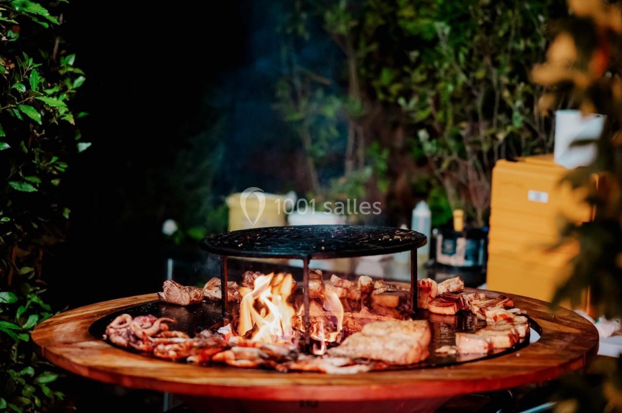 Viande et légumes grillant sur un barbecue rond avec un feu au centre, entouré de verdure en arrière-plan.