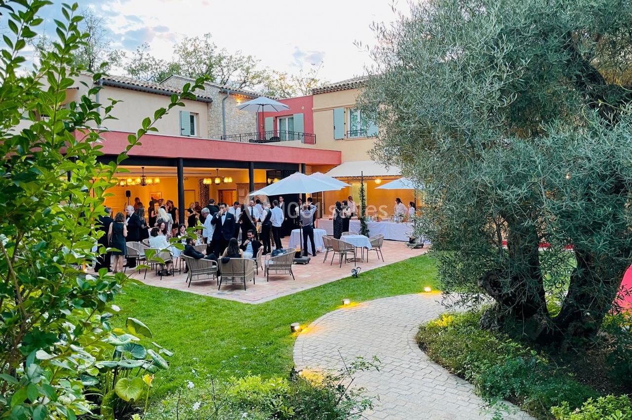Terrasse d'une maison avec des invités rassemblés autour de tables et parasols, entourée de verdure et d'arbres.