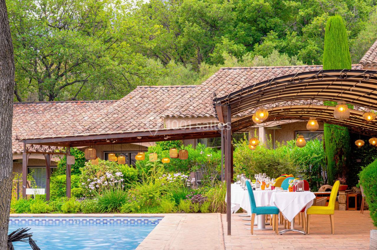 Terrasse avec table dressée près d'une piscine, entourée de végétation et éclairée par des lampes suspendues.