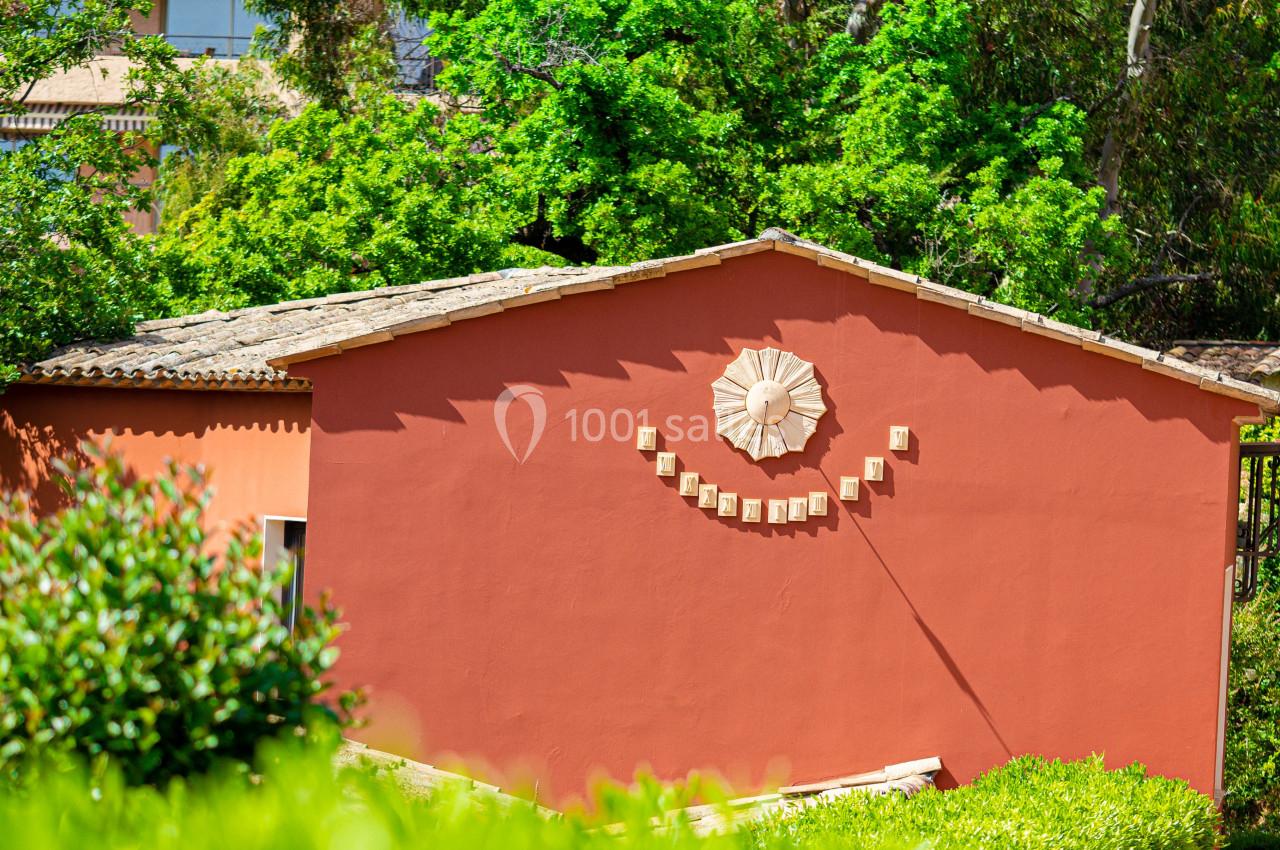 Façade d'une maison rouge ornée d'un décor en forme de sourire, entourée de verdure et d'arbres sous un ciel clair.