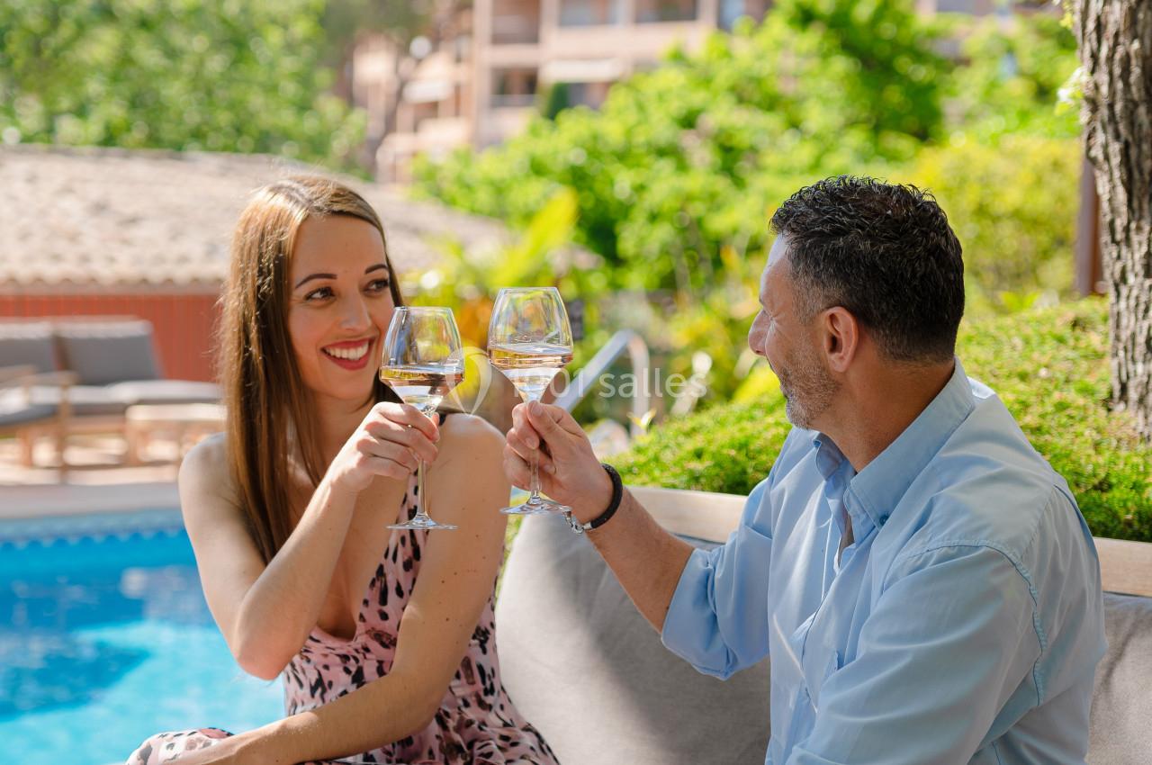 Un couple trinque avec des verres de vin blanc, assis près d'une piscine dans un cadre verdoyant et ensoleillé.