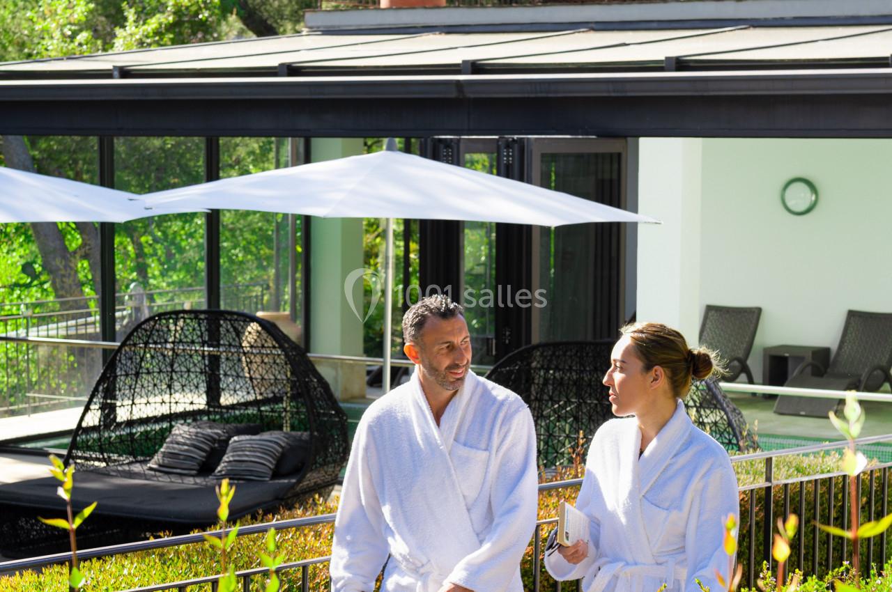 Deux personnes en peignoir discutent près d'une terrasse ensoleillée avec des chaises longues et des parasols.
