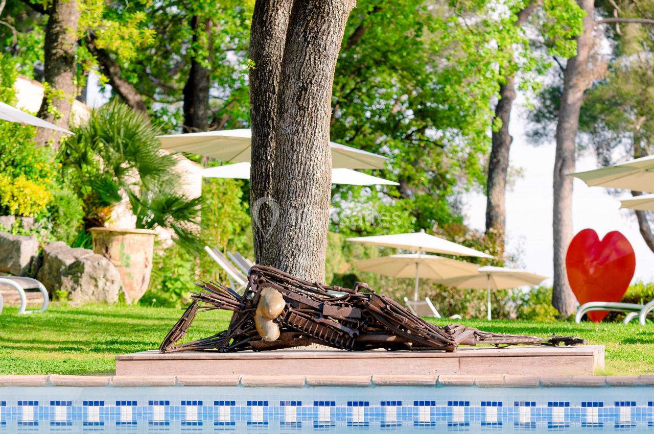Sculpture en bois représentant un insecte géant, placée près d'un arbre au bord d'une piscine entourée de verdure.