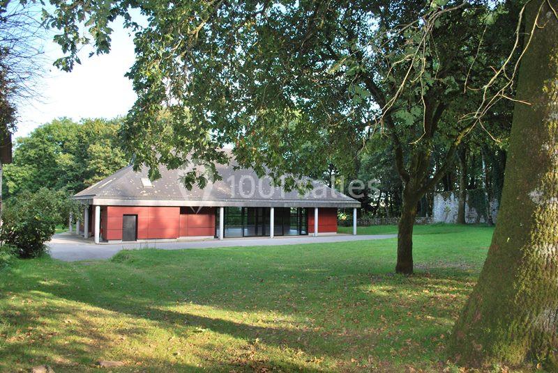 Location salle Plouay (Morbihan) - Domaine de Manehouarn #3 Bâtiment moderne avec façade rouge et grandes baies vitrées, entouré d'arbres et d'une pelouse verdoyante.