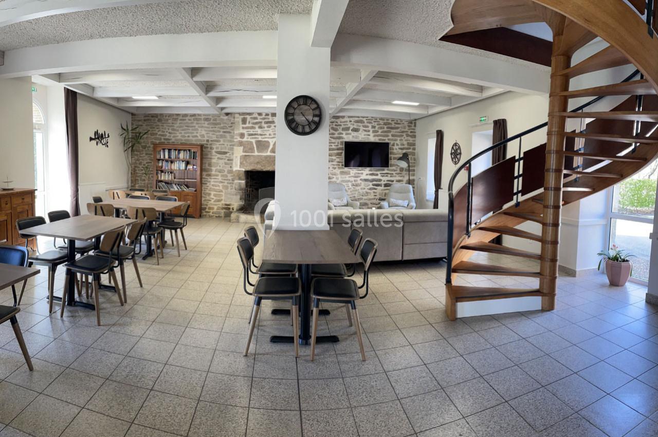 Location salle Plouay (Morbihan) - Domaine de Manehouarn #10 Salle lumineuse avec tables, chaises, canapé, escalier en bois, murs en pierre et grande horloge murale.
