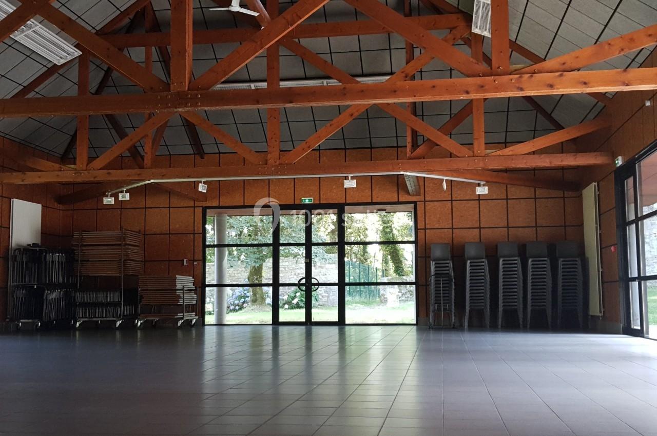 Location salle Plouay (Morbihan) - Domaine de Manehouarn #12 Salle spacieuse avec charpente en bois, grandes baies vitrées, chaises et tables empilées sur les côtés.