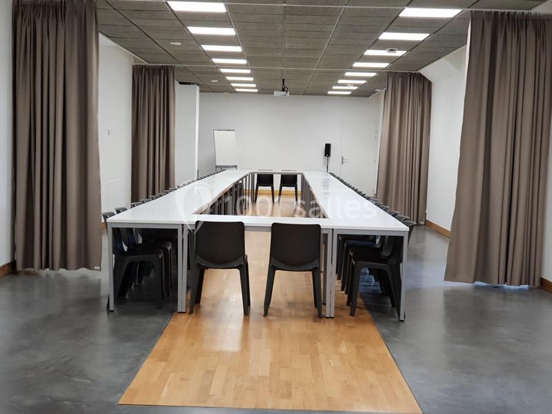 Salle de réunion avec tables disposées en U, chaises noires, rideaux beiges et tableau blanc en arrière-plan.