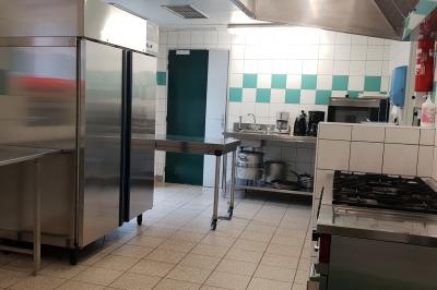 Cuisine professionnelle équipée avec réfrigérateur, plan de travail en inox, évier, plaques de cuisson et carrelage mural.