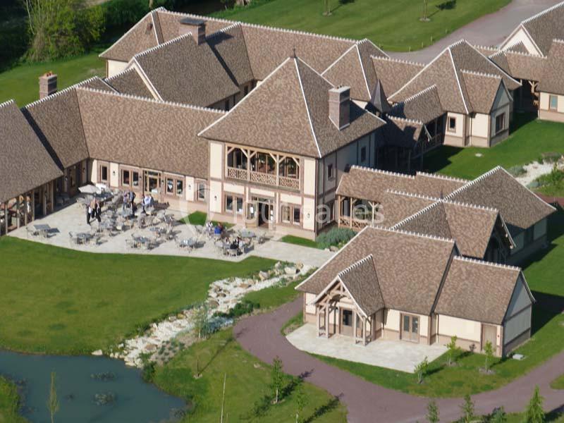 Vue aérienne d'un complexe de bâtiments en bois et pierre avec terrasses, entouré de pelouses et d'un étang.