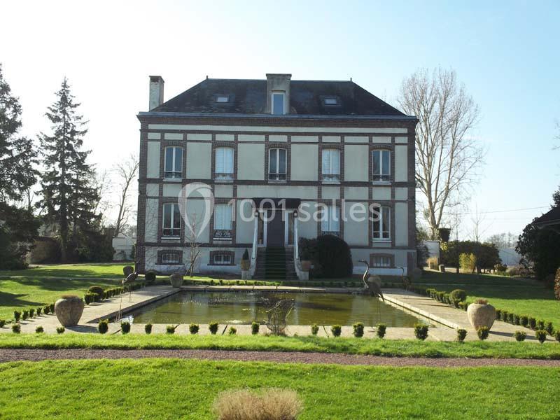 Grande maison en briques et pierre, entourée d'un jardin avec bassin central et allée bordée de buissons taillés.
