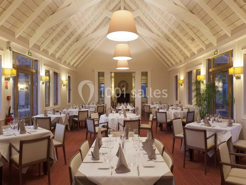 Salle de restaurant lumineuse avec tables dressées, chaises beige, grandes fenêtres et plafond voûté en bois blanc.