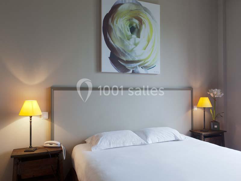 Lit double avec tête de lit beige, tables de chevet en bois, lampes allumées et tableau floral au mur.