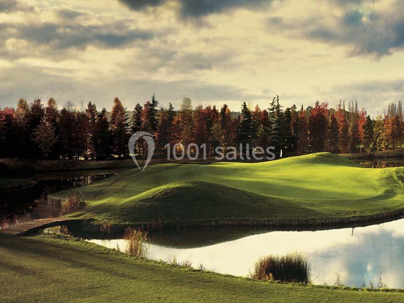 Paysage d'un terrain de golf entouré d'arbres aux couleurs automnales, avec un étang reflétant le ciel nuageux.
