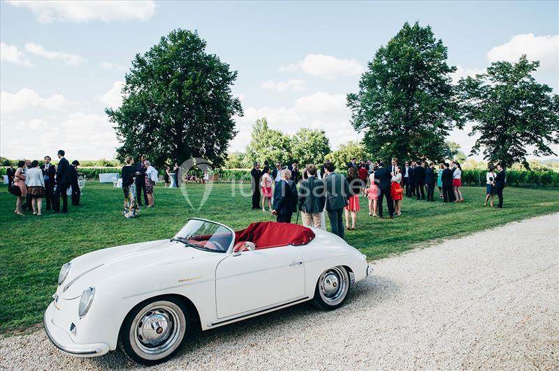 Voiture blanche décapotable garée sur une allée, avec des groupes de personnes discutant sur une pelouse en arrière-plan.