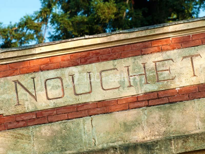 Inscription ’NOUCHET’ gravée sur une façade en pierre et briques, entourée de végétation en arrière-plan.