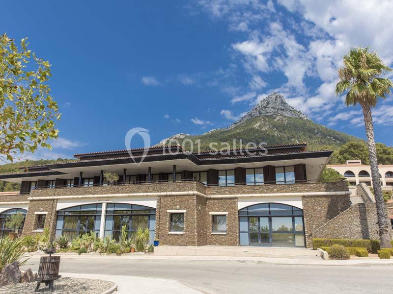 Bâtiment moderne en pierre avec grandes fenêtres, situé au pied d'une montagne sous un ciel partiellement nuageux.