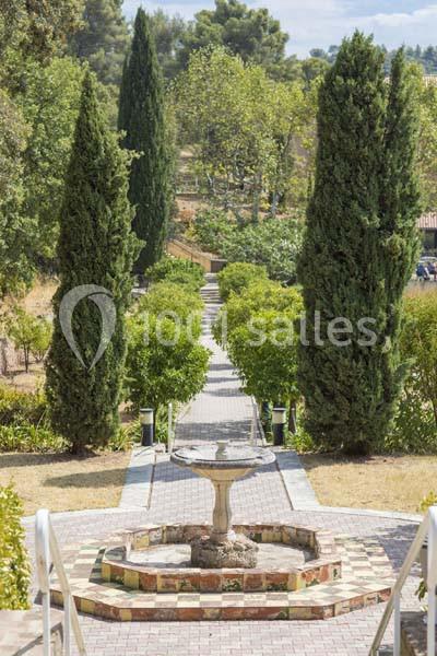 Allée pavée bordée de cyprès et d'arbustes, avec une fontaine centrale en pierre au premier plan.
