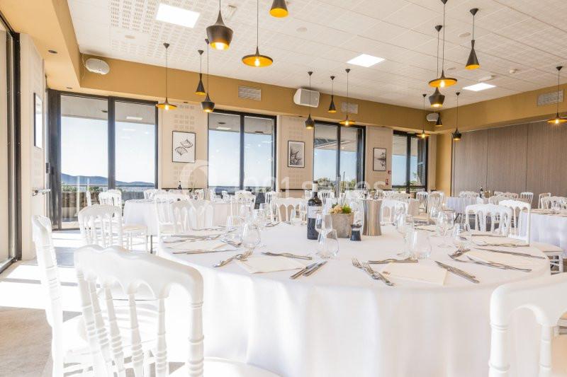 Salle lumineuse avec tables rondes dressées, chaises blanches et grandes baies vitrées offrant une vue dégagée.