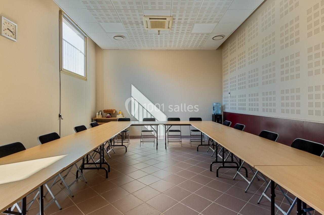 Salle de réunion lumineuse avec tables disposées en U, chaises noires, fenêtre, horloge murale et fontaine à eau.