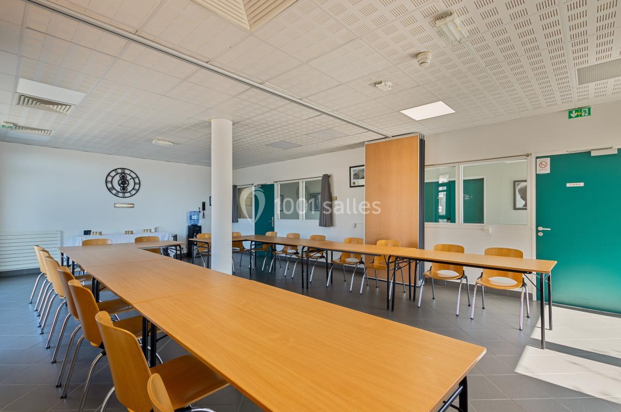 Salle de réunion lumineuse avec tables en bois disposées en U, chaises alignées et portes vertes en arrière-plan.
