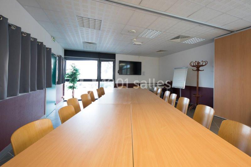 Salle de réunion avec une grande table en bois entourée de chaises, un écran mural et des rideaux gris.