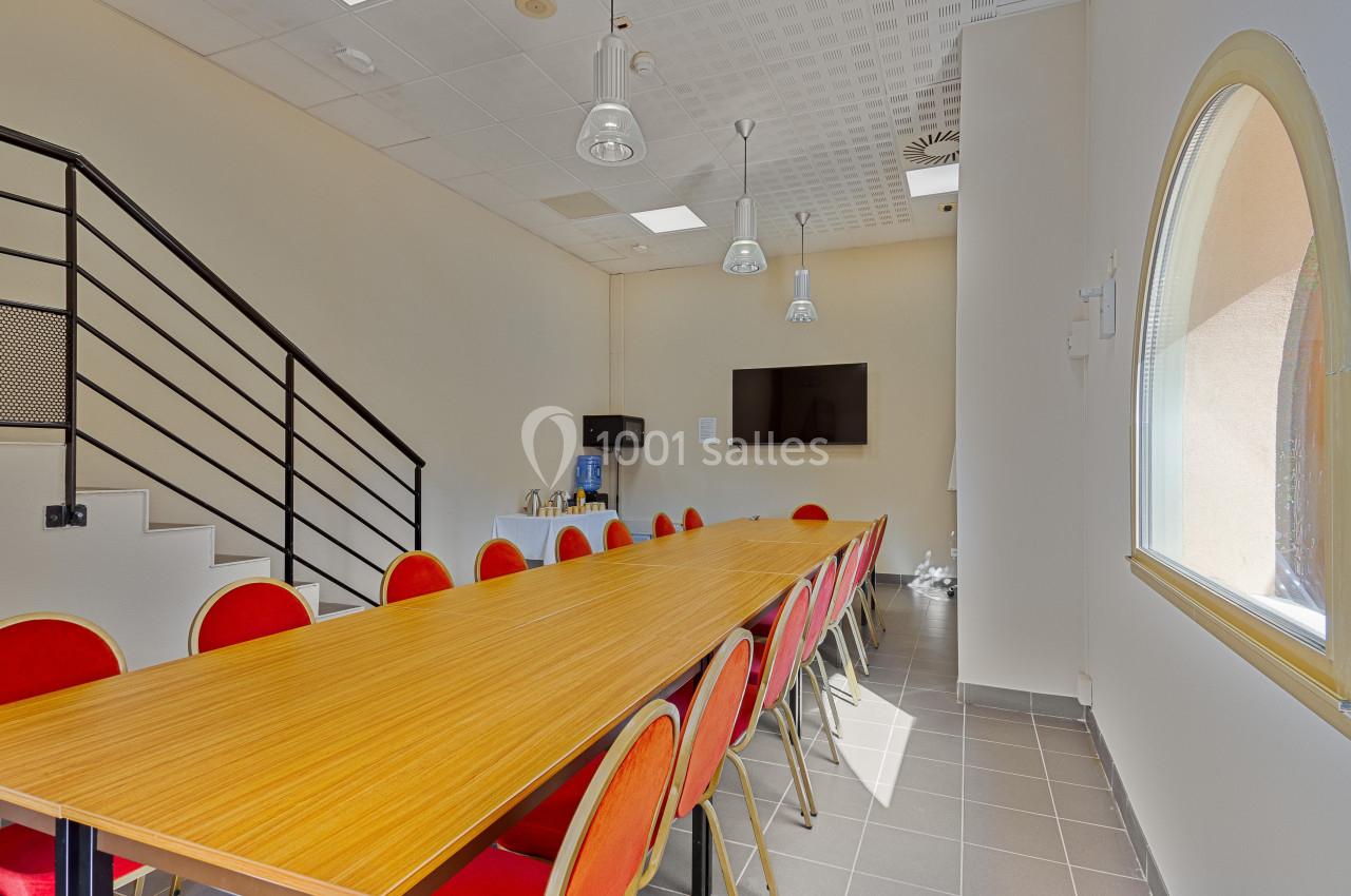Salle de réunion lumineuse avec une grande table en bois, des chaises rouges, un écran mural et un coin boissons.