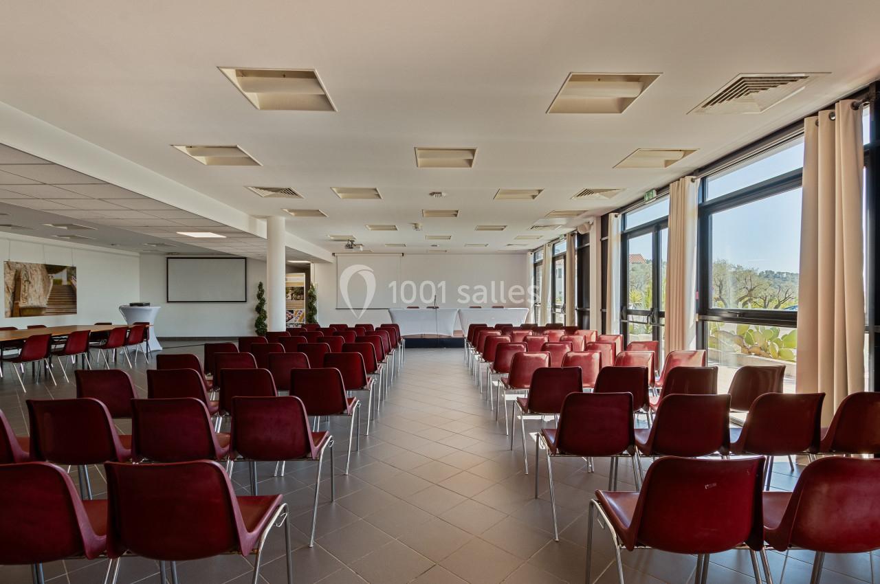 Salle de conférence lumineuse avec des rangées de chaises rouges, un tableau blanc et de grandes fenêtres.
