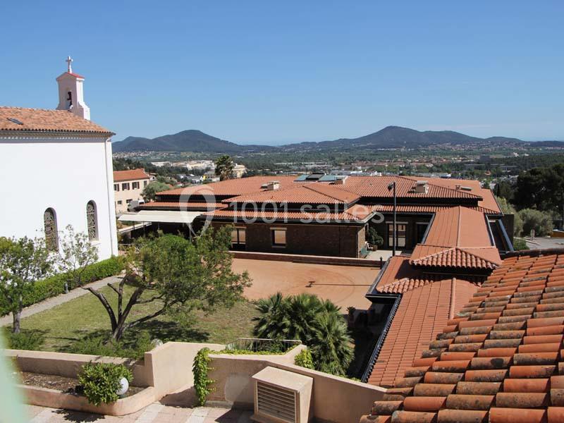 Vue d'un complexe de bâtiments avec toits en tuiles rouges, une église à gauche et des collines en arrière-plan.