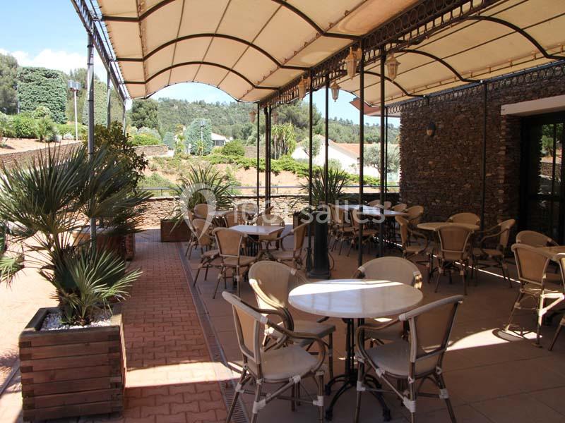 Terrasse extérieure ombragée avec tables et chaises, entourée de plantes et donnant sur un paysage verdoyant.