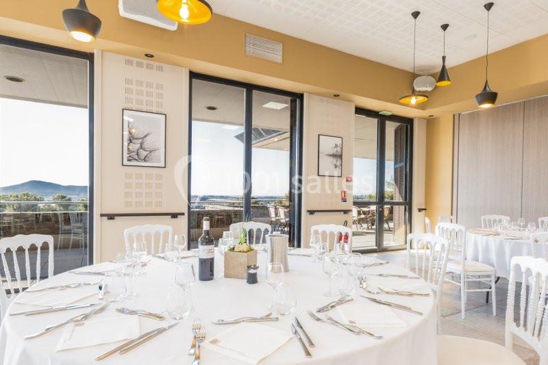 Salle lumineuse avec tables rondes dressées, chaises blanches, grandes baies vitrées et vue sur un paysage extérieur.