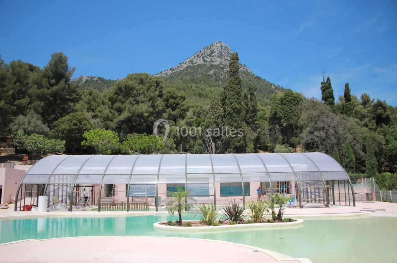 Piscine extérieure avec abri transparent, entourée de végétation et située au pied d'une colline boisée.
