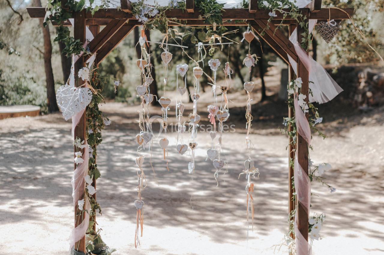 Arche en bois décorée de feuillage, rubans et suspensions, installée en extérieur sous des arbres.