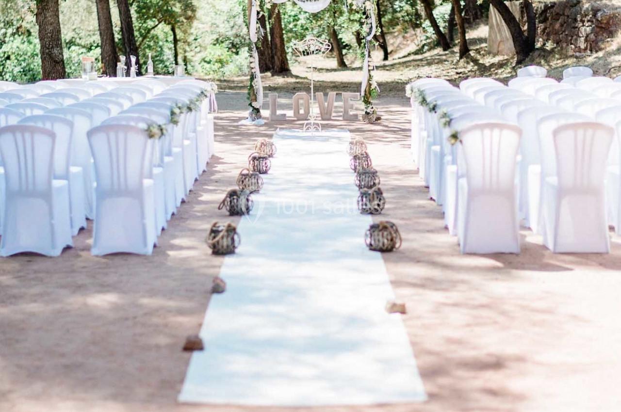 Allée centrale bordée de lanternes menant à une arche décorée de fleurs, entourée de chaises blanches en extérieur.