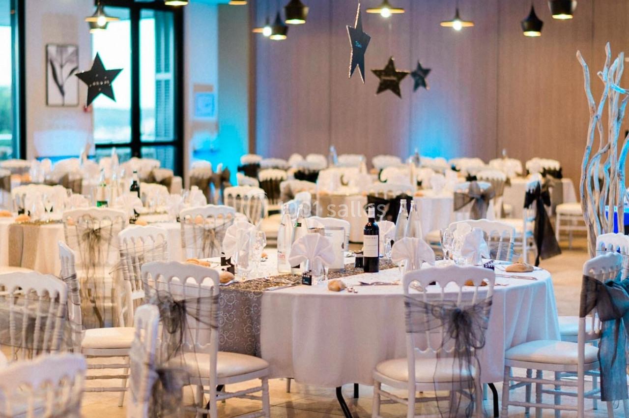 Salle de réception décorée avec des tables rondes dressées, chaises blanches et éclairage suspendu.