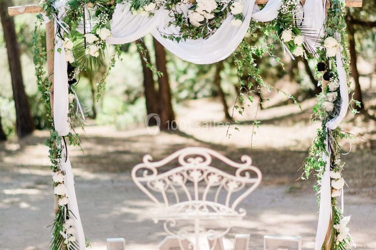 Arche de mariage décorée de fleurs, feuillages et tissus blancs, avec des lettres formant le mot ’LOVE’ au sol.