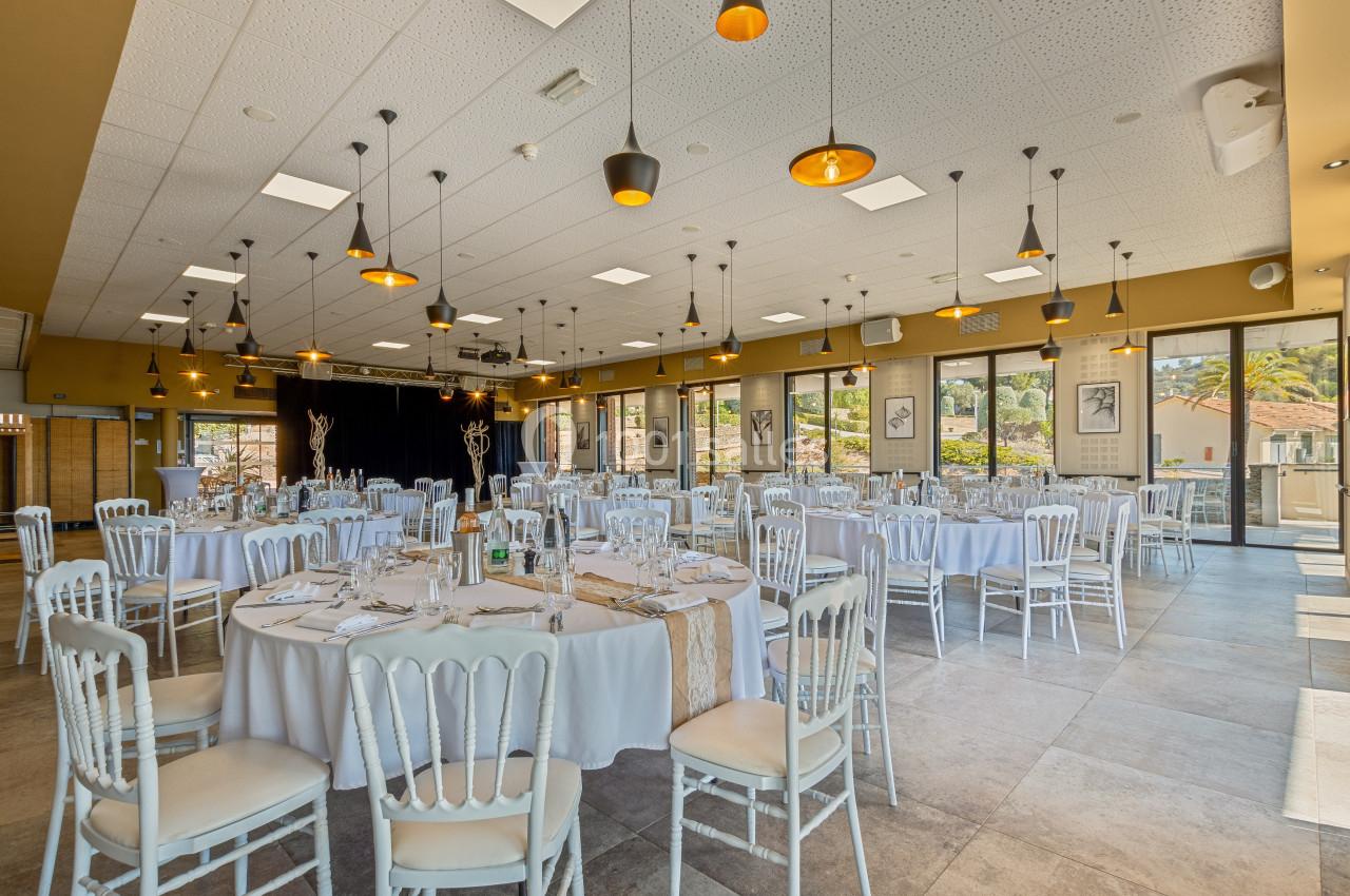 Salle de réception lumineuse avec tables rondes dressées, chaises blanches et grandes baies vitrées donnant sur l'extérieur.