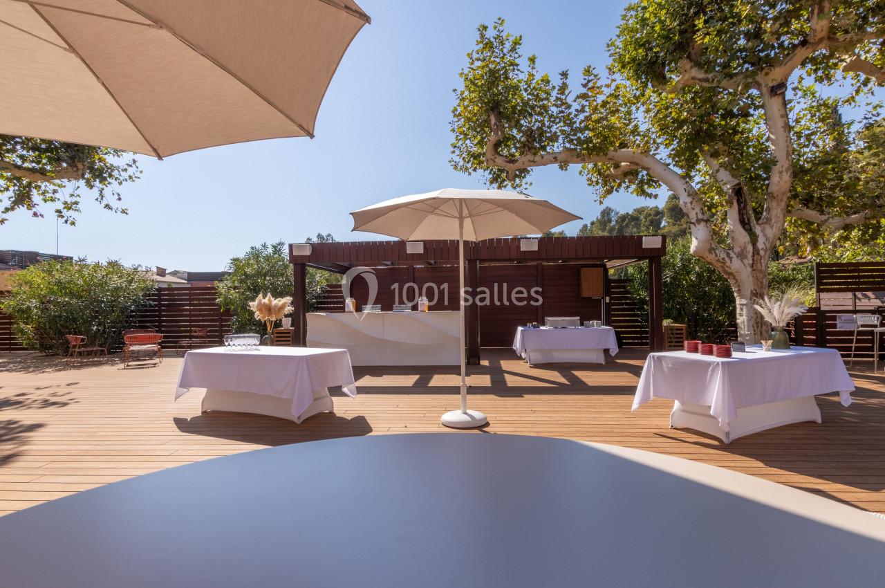 Terrasse ensoleillée avec tables dressées sous des parasols, entourée de végétation et de panneaux en bois.
