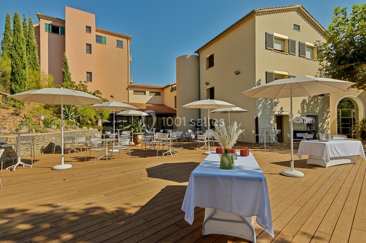 Terrasse en bois avec tables, chaises et parasols blancs, entourée de bâtiments colorés et de végétation.