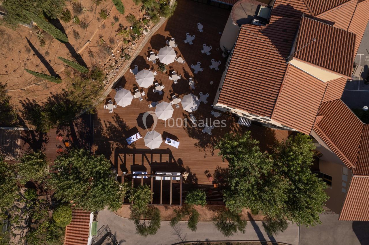 Vue aérienne d'une terrasse en bois avec des tables et parasols blancs, entourée de végétation et de bâtiments.