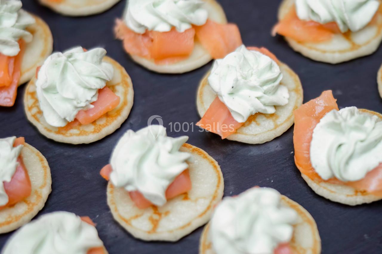 Petits blinis garnis de crème fouettée et de tranches de saumon fumé, disposés sur une surface sombre.