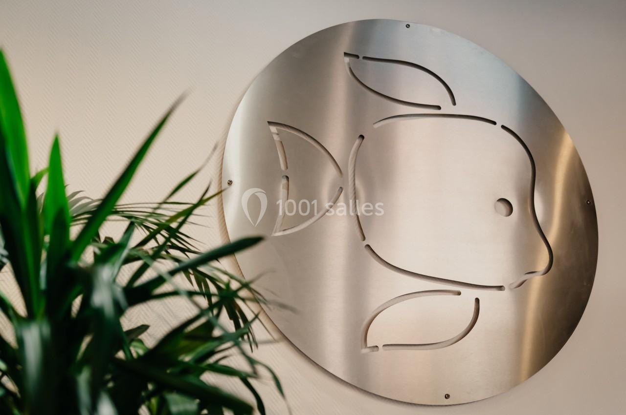 Location salle La Mulatière (Rhône) - Aquarium de Lyon #21 Plaque métallique ronde avec un motif de poisson découpé, accrochée à un mur clair, partiellement cachée par une plante…